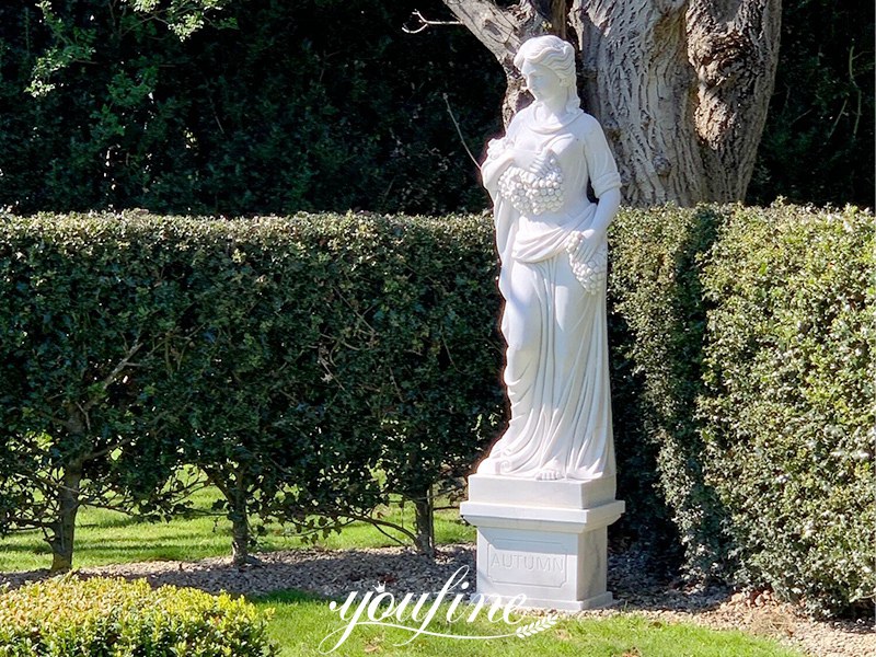 White Four Seasons female statue positioned beside a hedge in a garden
