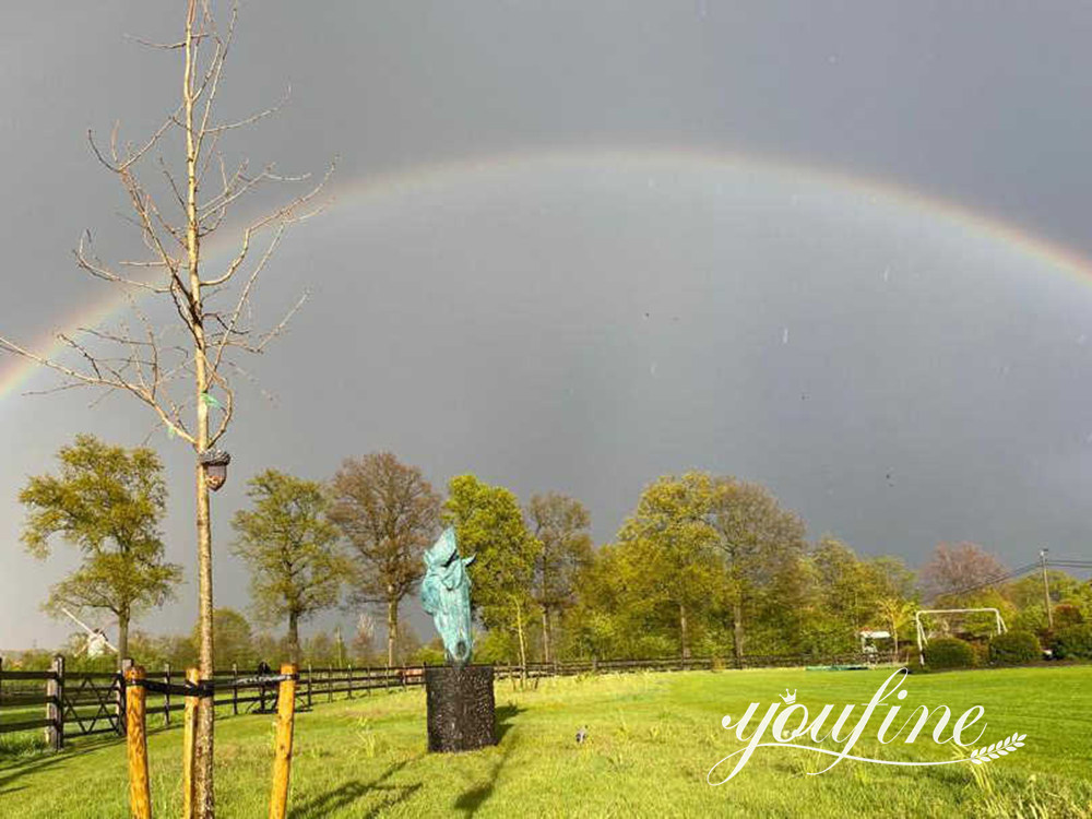 Eine grüne Pferdekopfskulptur auf einem dunklen Sockel im Garten eines belgischen Kunden, dahinter ein Regenbogen.